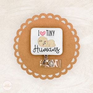 May include: A white badge reel with the text "I ♥ TINY Humans" and an embroidered baby design. The badge is attached to a brown scalloped circle. The reel has a clear plastic strap and a metal clip.