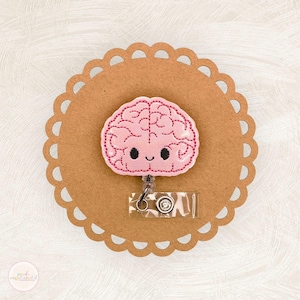 May include: A pink brain-shaped badge reel with a smiling face, stitched with pink thread. The badge reel is attached to a clear plastic holder and a metal clip. The badge reel is on a brown, scalloped-edge paper.
