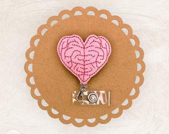 Op de afbeelding: Een roze vilten badge reel in de vorm van een hersen, gestikt met donkerder roze draad. De badge reel is bevestigd aan een doorzichtige plastic houder met een metalen clip. De badge staat op een bruine achtergrond met geschulpte rand.
