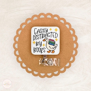 May include: A white badge reel with the text "Easily Distracted by Books" and an embroidered duck carrying books. The badge is on a brown, scalloped-edge background. A clear plastic reel and metal clip are attached.