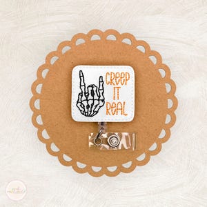 May include: A white badge reel with the text "Creep It Real" in orange, featuring a black skeleton hand gesture. The badge is attached to a clear plastic reel with a metal clip, set on a scalloped brown background.