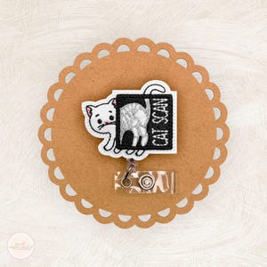 May include: A white cat-themed badge reel with the text "CAT SCAN" on a black background. The badge reel is attached to a brown, scalloped-edge circle.