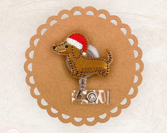 May include: A brown dachshund dog badge reel with a red and white Santa hat. The badge reel is attached to a clear plastic retractable reel with a metal clip. The badge reel is on a brown scalloped circle.
