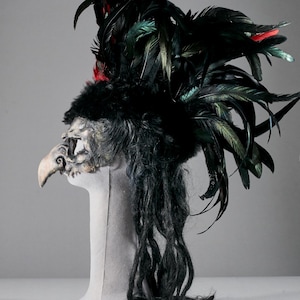 May include: A theatrical mask featuring a bird-like design. The mask is primarily black with accents of red and green feathers. It has a long, beak-like nose and is mounted on a white mannequin head. Long, black, dreadlock-like strands hang from the mask.