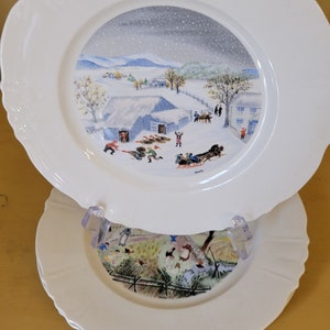 Puede incluir: Dos platos de cer&aacute;mica blanca con una escena de invierno pintada en el plato superior. La escena representa un paisaje nevado con una granja, &aacute;rboles y personas desliz&aacute;ndose en trineo. El plato inferior tiene un paisaje verde y marr&oacute;n con una valla y &aacute;rboles.