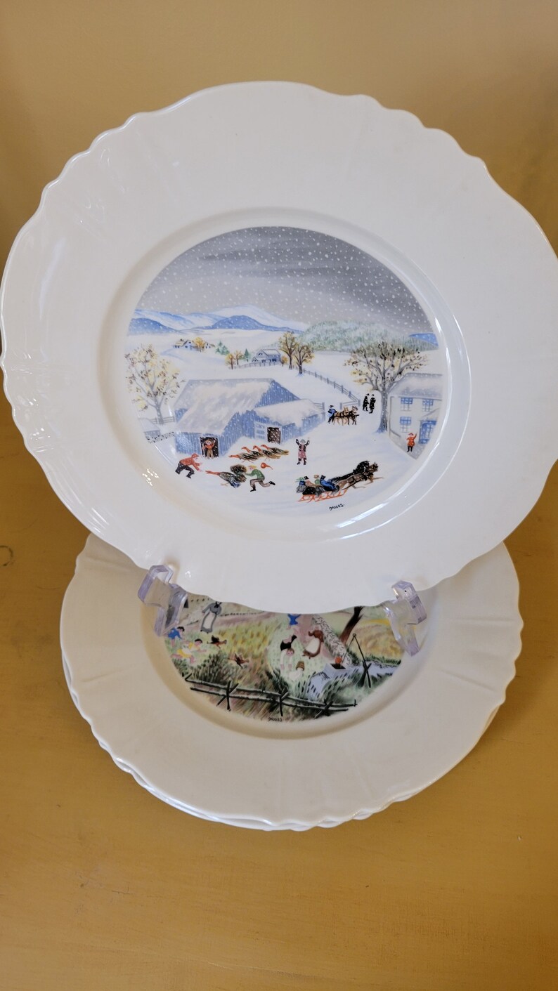 Puede incluir: Dos platos de cer&aacute;mica blanca con una escena de invierno pintada en el plato superior. La escena representa un paisaje nevado con una granja, &aacute;rboles y personas desliz&aacute;ndose en trineo. El plato inferior tiene un paisaje verde y marr&oacute;n con una valla y &aacute;rboles.