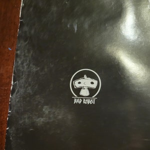 Puede incluir: Un p&oacute;ster en blanco y negro con el logotipo de Bad Robot. El logotipo es un robot blanco con un c&iacute;rculo a su alrededor. El texto "Bad Robot" est&aacute; debajo del robot. El p&oacute;ster tambi&eacute;n tiene el texto "PRINTED IN U.S.A." en la parte inferior.