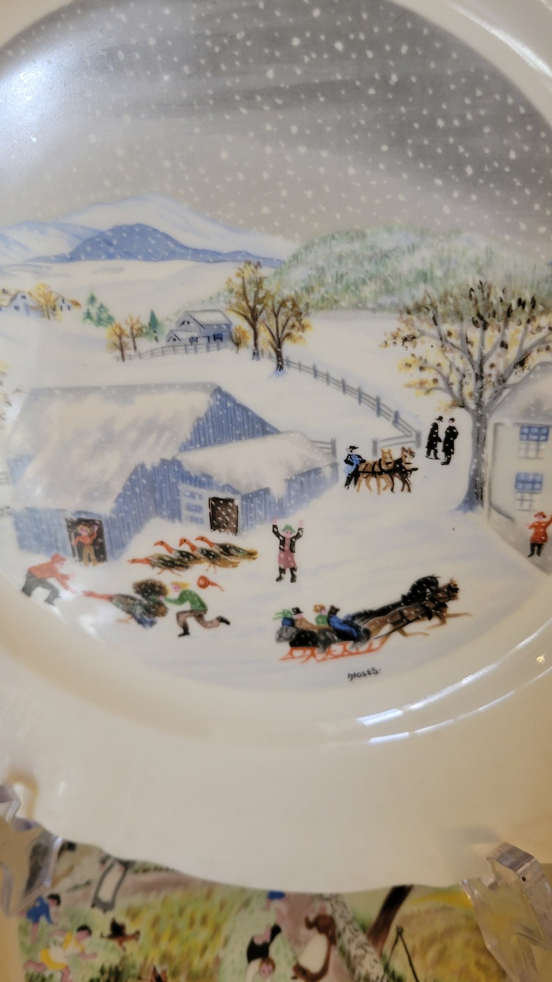 Puede incluir: Un plato de cer&aacute;mica vintage con una escena de invierno. El plato presenta un paisaje nevado con una granja, &aacute;rboles y personas. La escena est&aacute; pintada en un estilo caprichoso con colores brillantes. El plato est&aacute; firmado "Moses".