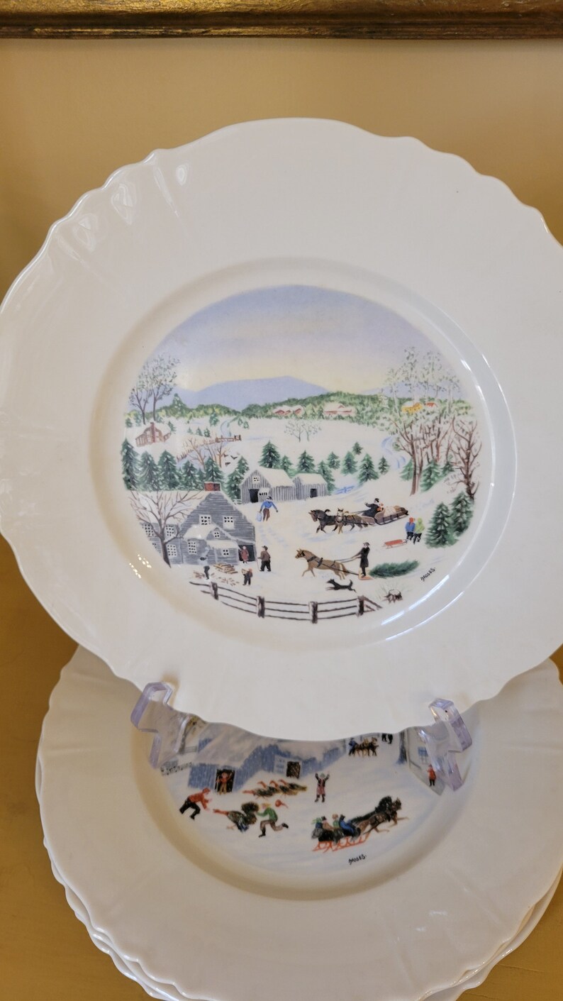 Puede incluir: Un juego de cuatro platos de cer&aacute;mica blanca con una escena de invierno pintada en ellos. La escena representa un paisaje nevado con casas, &aacute;rboles y personas. Los platos tienen un borde festoneado y son perfectos para una cena navide&ntilde;a.