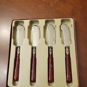 Puede incluir: Juego de cuatro cuchillos de mantequilla de plata con mangos rojos y dorados en una caja de pl&aacute;stico transparente. Los cuchillos est&aacute;n en fila y los mangos est&aacute;n todos orientados en la misma direcci&oacute;n.
