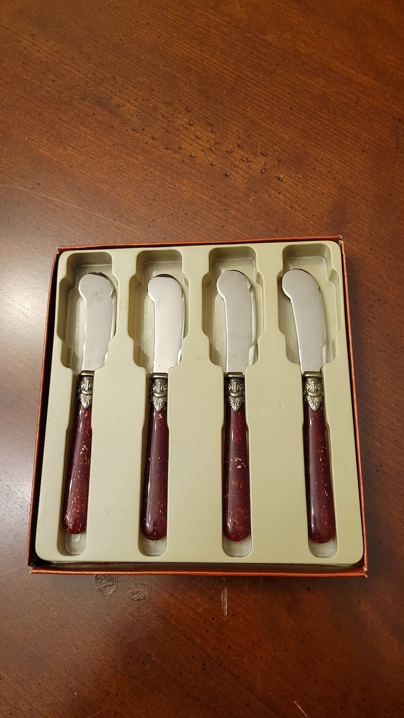 Puede incluir: Juego de cuatro cuchillos de mantequilla de plata con mangos rojos y dorados en una caja de pl&aacute;stico transparente. Los cuchillos est&aacute;n en fila y los mangos est&aacute;n todos orientados en la misma direcci&oacute;n.