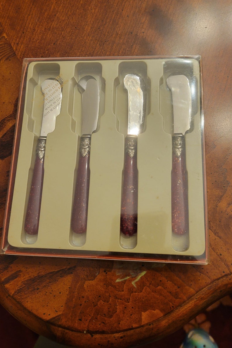 Puede incluir: Juego de cuatro cuchillos de mantequilla de plata con mangos burdeos. Los cuchillos est&aacute;n en una caja de pl&aacute;stico transparente con fondo blanco. La caja tiene el texto "Stainless Dishwasher Safe Made in Italy Dish Washer Safe" impreso en ella.