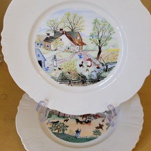 Puede incluir: Un juego de tres platos de cer&aacute;mica blanca con borde festoneado. El plato superior presenta una escena pintada a mano de una casa de campo con un c&eacute;sped verde, &aacute;rboles y personas. Los dos platos inferiores tienen un dise&ntilde;o similar con una escena diferente.