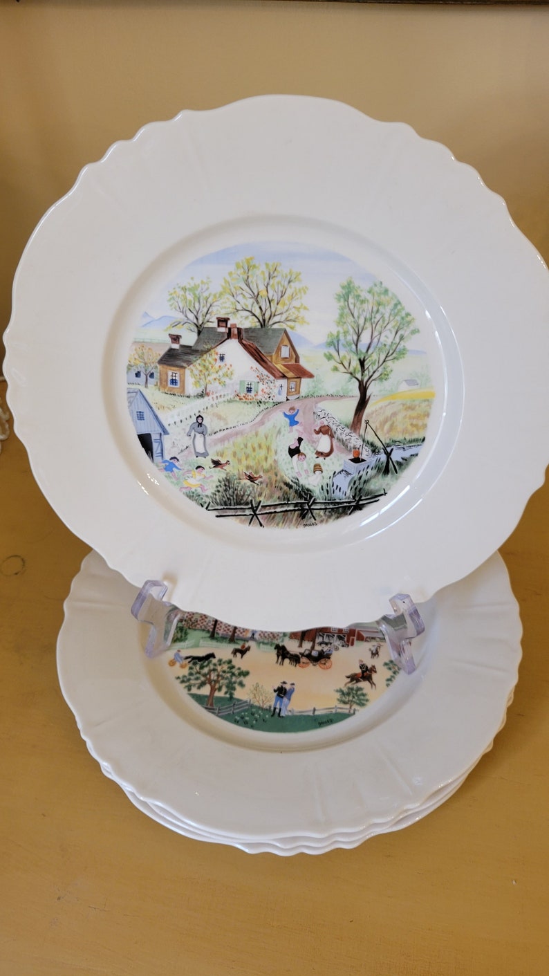 Puede incluir: Un juego de tres platos de cer&aacute;mica blanca con borde festoneado. El plato superior presenta una escena pintada a mano de una casa de campo con un c&eacute;sped verde, &aacute;rboles y personas. Los dos platos inferiores tienen un dise&ntilde;o similar con una escena diferente.