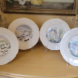 Puede incluir: Cuatro platos de cer&aacute;mica blanca con bordes decorativos, cada uno con una escena de invierno diferente pintada en estilo acuarela. Los platos se muestran sobre una superficie de madera.