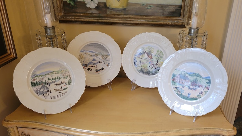 Puede incluir: Cuatro platos de cer&aacute;mica blanca con bordes decorativos, cada uno con una escena de invierno diferente pintada en estilo acuarela. Los platos se muestran sobre una superficie de madera.