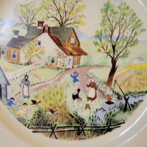 Puede incluir: Un plato de cer&aacute;mica blanco con una escena pintada colorida de una granja rural con un pozo y ni&ntilde;os jugando. El plato est&aacute; firmado "Moses".