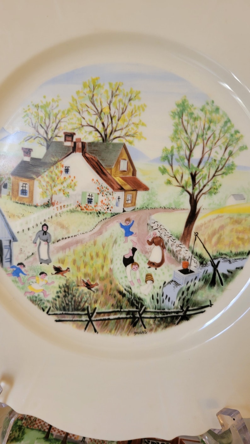 Puede incluir: Un plato de cer&aacute;mica blanco con una escena pintada colorida de una granja rural con un pozo y ni&ntilde;os jugando. El plato est&aacute; firmado "Moses".