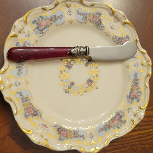 Puede incluir: Un plato de porcelana blanca con un borde dorado y un patr&oacute;n floral. Un cuchillo de mantequilla con mango rojo est&aacute; encima del plato.