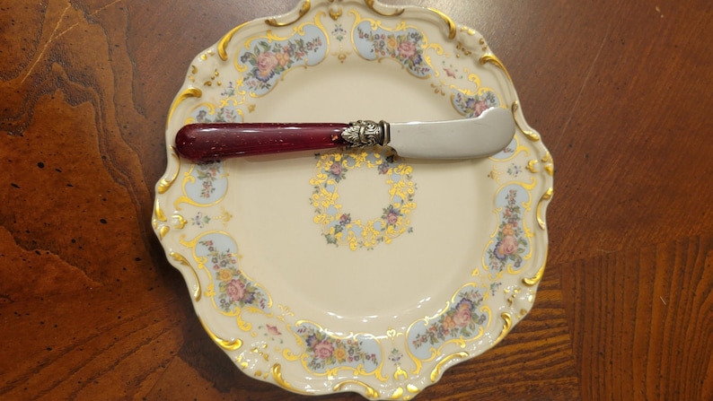 Puede incluir: Un plato de porcelana blanca con un borde dorado y un patr&oacute;n floral. Un cuchillo de mantequilla con mango rojo est&aacute; encima del plato.