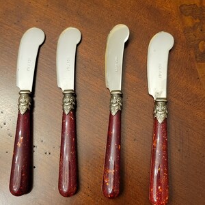 Puede incluir: Cuatro cuchillos de mantequilla de plata con mangos rojos y dorados. Los cuchillos tienen las palabras "Made in Italy" grabadas en las hojas.