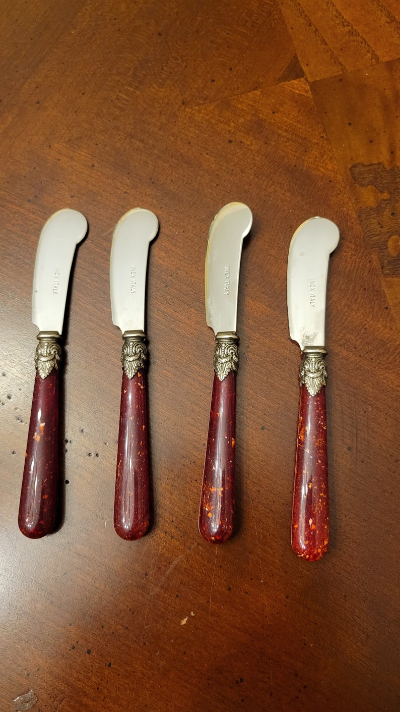 Puede incluir: Cuatro cuchillos de mantequilla de plata con mangos rojos y dorados. Los cuchillos tienen las palabras "Made in Italy" grabadas en las hojas.