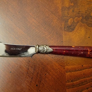 Puede incluir: Un cuchillo de mantequilla de plata con un mango rojo y dorado moteado. El cuchillo tiene las palabras "Inox Italy" grabadas en la hoja.