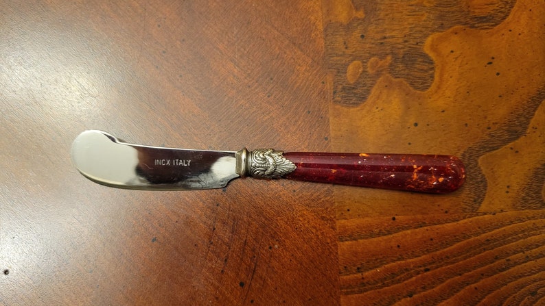 Puede incluir: Un cuchillo de mantequilla de plata con un mango rojo y dorado moteado. El cuchillo tiene las palabras "Inox Italy" grabadas en la hoja.