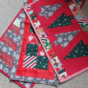 May include: A quilted table runner featuring a patchwork design with red, green, and grey fabrics. The runner displays appliquéd Christmas tree shapes, snowflake patterns, and festive motifs including Santa Claus, all bordered by a red and white gingham trim.