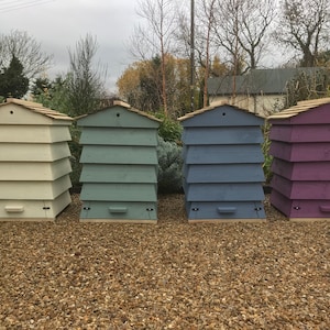 May include: Four wooden beehives painted in different colours. The beehives are stacked in a row, from left to right: white, light blue, dark blue, and purple.