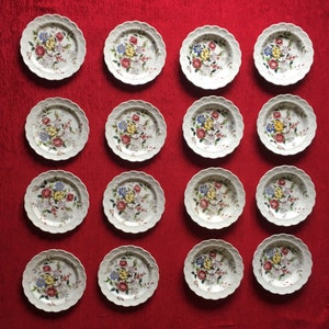 May include: A collection of 15 white ceramic plates with floral designs in shades of pink, blue, yellow, and green. The plates have a scalloped edge and are arranged in a grid pattern.