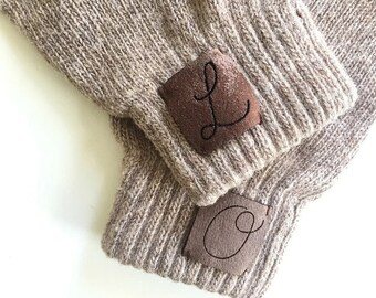 Puede incluir: Dos pares de guantes sin dedos de punto marrón con parches de cuero marrón. Los parches tienen las letras "L" y "O" bordadas en hilo negro.
