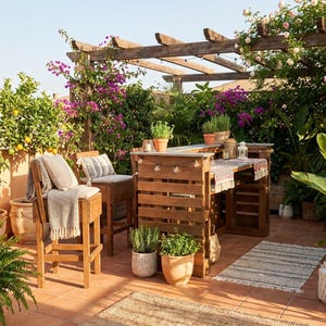 Op de afbeelding: Buitenterras met een houten bar, krukken en een pergola. De bar is gemaakt van houten pallets. Potplanten en bloemen omringen de ruimte en creëren een ontspannen sfeer. Een klein tapijt en kussens zorgen voor comfort.