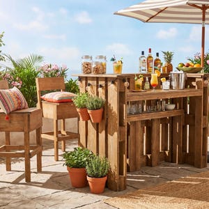 Puede incluir: Barra de madera al aire libre con sombrilla a rayas, bebidas y taburetes. La barra tiene estantes y está abastecida con botellas, vasos y una cubitera de metal. Dos taburetes de madera con cojines están a la izquierda. Plantas y hierbas en macetas.