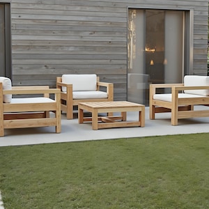 Peut inclure: Quatre chaises en bois avec des coussins blancs et une table basse en bois assortie. Les chaises et la table sont disposées en carré sur une terrasse avec un mur en bois gris en arrière-plan.