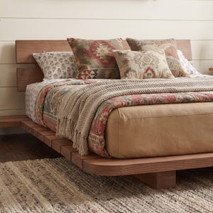 May include: A low-profile wooden platform bed frame with a horizontal slat headboard. The bed is dressed with a tan leather-covered mattress, a patterned quilt in shades of red and green, a beige knit throw blanket, and several decorative pillows.