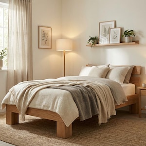 May include: A light-filled bedroom with a wooden bed frame, a beige comforter, and a gray blanket. The room features a jute rug, a bedside table with a lamp, and a window with sheer curtains. Decorative framed botanical prints and potted plants add a touch of nature.