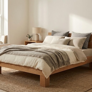 May include: A wooden bed with a neutral duvet, pillows, and a grey throw. The bed is in a bright room with a window and a woven rug. A lamp and side table are visible. The bed frame is approximately 200cm long.