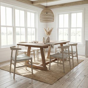 May include: A light brown wooden dining table with six matching chairs. The table is set on a woven jute rug. A woven pendant light hangs above the table. The room has white walls and large windows.
