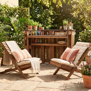 May include: Two wooden lounge chairs with cream cushions and striped pillows sit on a stone patio. A wooden bar with shelves and a "WELCOME" sign is in the background, surrounded by potted plants and greenery. A cozy outdoor setting.
