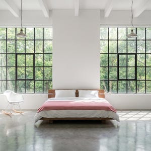 Könnte beinhalten: Ein modernes Schlafzimmer mit einem niedrigen Bettrahmen aus Holz, weißer Bettwäsche und einer rot-weiß karierten Decke. Große Fenster mit schwarzen Rahmen bieten einen Blick auf üppige grüne Bäume. Ein weißer Schaukelstuhl steht in der Ecke, und zwei Pendelleuchten hängen von der Decke.