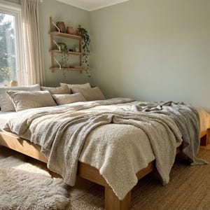 May include: A light wood bed frame with a neutral-toned comforter and pillows. A fluffy, cream-colored blanket is draped over the bed. A woven rug and a paper lantern lamp add to the cozy bedroom decor.