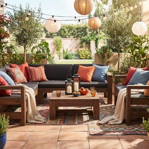 May include: Outdoor seating arrangement featuring a dark grey sectional sofa, wooden coffee table, and armchairs. Cushions and throws in various colours. Lanterns and string lights create a relaxed atmosphere.