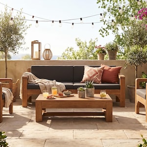 May include: Outdoor patio furniture set with a wooden frame, featuring a sofa, two armchairs, and a coffee table. The set includes dark gray cushions, decorative pillows, and a beige throw blanket. The scene is set on a stone patio with plants and string lights.