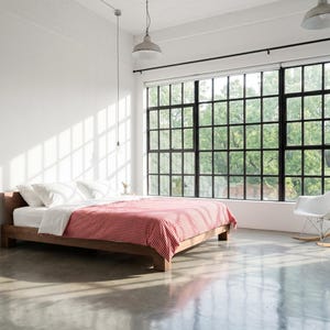 Könnte beinhalten: Ein modernes Schlafzimmer mit einem Bettrahmen aus Holz, weißer Bettwäsche und einer rot-weiß karierten Decke. Der Raum verfügt über ein großes Fenster mit schwarzen Rahmen, einen weißen Schaukelstuhl und Hängeleuchten. Der Boden ist polierter Beton.
