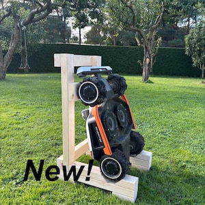 Pode incluir: Um robô cortador de grama laranja e preto é exibido em um suporte de madeira em um jardim gramado. A palavra "New!" está escrita em texto preto no canto inferior esquerdo. O cortador de grama tem rodas pretas grandes e um topo prateado.