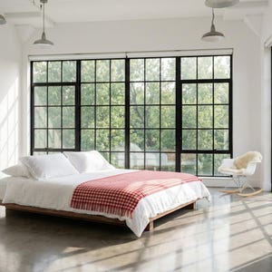 Peut inclure: Une chambre moderne avec un cadre de lit bas en bois. Le lit est recouvert de draps blancs et d'une couverture à carreaux rouges et blancs. De grandes fenêtres offrent une vue sur la verdure. Un fauteuil à bascule blanc avec une peau de mouton est à proximité.