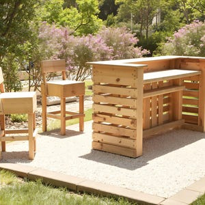 Peut inclure: Un bar extérieur en bois et deux chaises assorties. Le bar a un comptoir et une étagère, construits en bois clair. Les chaises ont un design simple. L'ensemble est posé sur une surface de gravier, entouré de verdure.