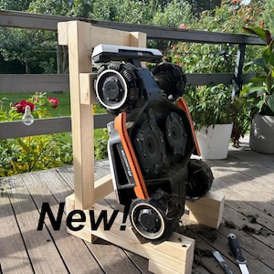 May include: A robotic lawn mower, black and orange, is mounted on a wooden stand. The mower has large, textured wheels and is labeled "New!" in white text. The background includes a wooden deck and greenery.