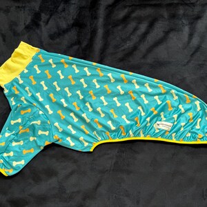 May include: A turquoise pet shirt featuring a repeating pattern of yellow and white dog bones. The garment has a bright yellow collar and yellow trim along the sleeve and bottom hem. A small white rectangular label is attached to the side.